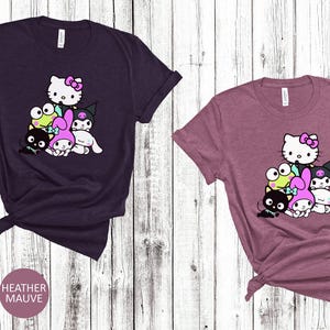 K&ouml;nnte beinhalten: Zwei T-Shirts mit einem Grafikdruck einer Gruppe von Comicfiguren. Die Figuren sind Hello Kitty, My Melody, Kuromi und ein Frosch. Die T-Shirts sind in den Farben Heather Maroon und Heather Mauve erh&auml;ltlich.
