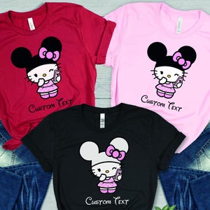 Pu&ograve; includere: T-shirt rosse, rosa e nere con un design di personaggi dei cartoni animati. Ogni maglietta presenta un personaggio con un fiocco, che tiene in mano un telefono e indossa un vestito. Il testo "Custom Text" &egrave; sotto il personaggio. Jeans blu sullo sfondo.