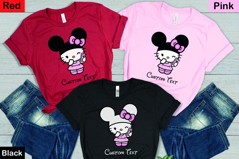 Pu&ograve; includere: T-shirt rosse, rosa e nere con un design di personaggi dei cartoni animati. Ogni maglietta presenta un personaggio con un fiocco, che tiene in mano un telefono e indossa un vestito. Il testo "Custom Text" &egrave; sotto il personaggio. Jeans blu sullo sfondo.