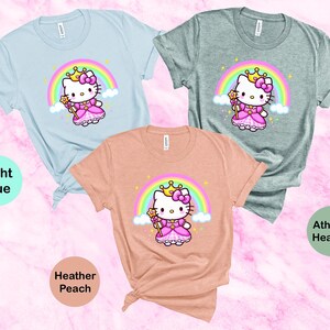 May include: Three t-shirts in light blue, athletic heather, and heather peach. Each shirt features a cartoon princess cat design with a rainbow, crown, and wand. The text "Light Blue", "Athletic Heather", and "Heather Peach" are also visible.