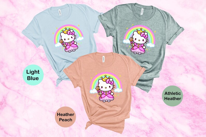May include: Three t-shirts in light blue, athletic heather, and heather peach. Each shirt features a cartoon princess cat design with a rainbow, crown, and wand. The text "Light Blue", "Athletic Heather", and "Heather Peach" are also visible.