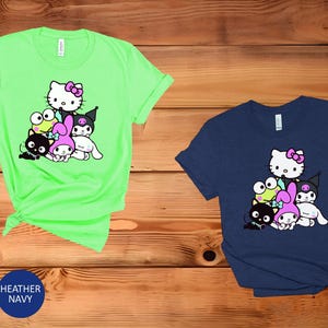 K&ouml;nnte beinhalten: Zwei T-Shirts mit einem grafischen Druck einer Gruppe von Comicfiguren. Das Shirt links ist hellgr&uuml;n und das Shirt rechts dunkelblau. Zu den Figuren geh&ouml;ren Hello Kitty, My Melody, Kuromi und ein Frosch.