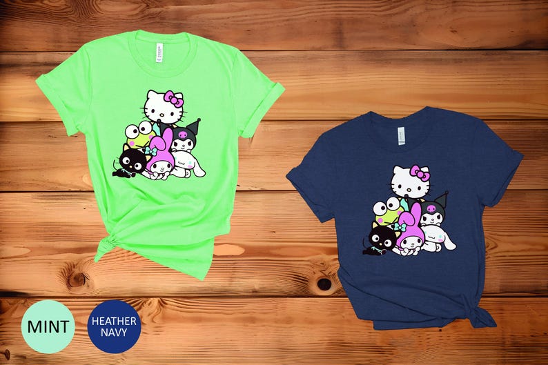 K&ouml;nnte beinhalten: Zwei T-Shirts mit einem grafischen Druck einer Gruppe von Comicfiguren. Das Shirt links ist hellgr&uuml;n und das Shirt rechts dunkelblau. Zu den Figuren geh&ouml;ren Hello Kitty, My Melody, Kuromi und ein Frosch.