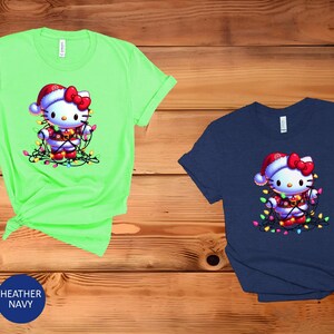 May include: Two short-sleeved t-shirts, one mint green and one navy blue, featuring a cartoon character wearing a Santa hat and wrapped in Christmas lights. The character has a red bow and is wearing a festive outfit.