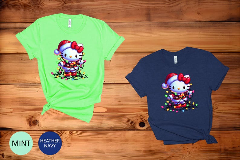 May include: Two short-sleeved t-shirts, one mint green and one navy blue, featuring a cartoon character wearing a Santa hat and wrapped in Christmas lights. The character has a red bow and is wearing a festive outfit.