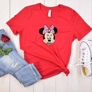 May include: Red t-shirt with a graphic of Minnie Mouse wearing a pink bow with white polka dots. The shirt is laid out on a white wooden surface with blue jeans, a red rose, and white sneakers.