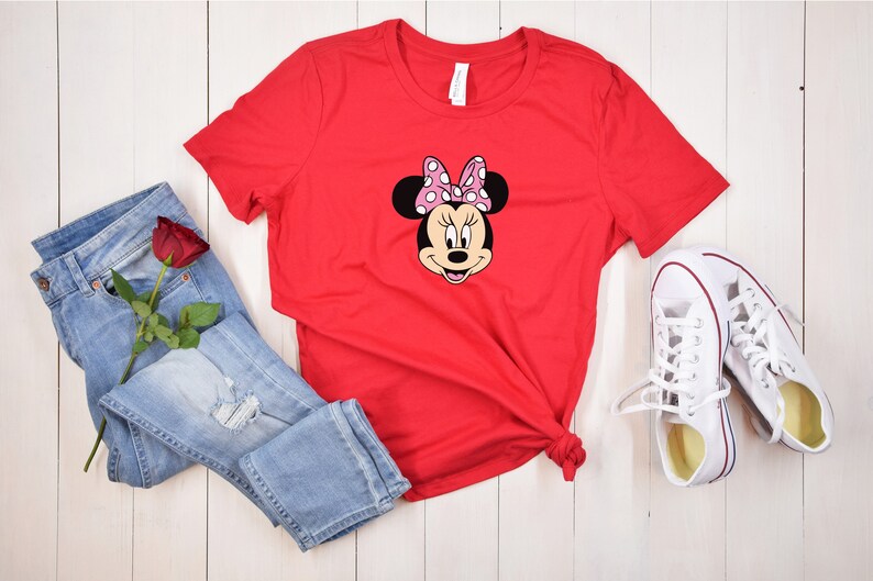 May include: Red t-shirt with a graphic of Minnie Mouse wearing a pink bow with white polka dots. The shirt is laid out on a white wooden surface with blue jeans, a red rose, and white sneakers.