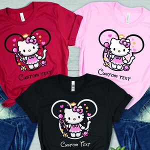 May include: Three t-shirts with a pink Hello Kitty character wearing a crown and holding a wand. The shirts are red, pink, and black. The text "Custom Text" is printed on each shirt.