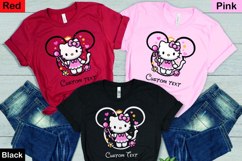 May include: Three t-shirts with a pink Hello Kitty character wearing a crown and holding a wand. The shirts are red, pink, and black. The text "Custom Text" is printed on each shirt.