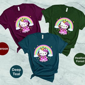 May include: Three short-sleeved t-shirts in maroon, deep teal, and heather forest green. Each shirt features a cartoon princess cat design with a rainbow, crown, and wand. The shirts are displayed on a light gray surface.