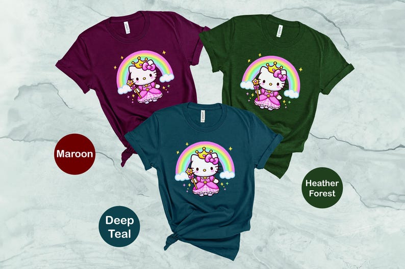 May include: Three short-sleeved t-shirts in maroon, deep teal, and heather forest green. Each shirt features a cartoon princess cat design with a rainbow, crown, and wand. The shirts are displayed on a light gray surface.