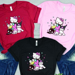 Pu&ograve; includere: Tre magliette con una stampa grafica di Hello Kitty e i suoi amici. Le magliette sono rossa, rosa e nera. La grafica presenta Hello Kitty, My Melody, Badtz-Maru e Keroppi.