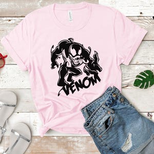 May include: A pink t-shirt with a black graphic of Venom, a fictional superhero, with the word "VENOM" printed below the graphic.