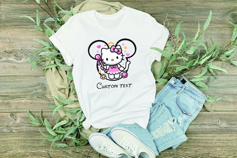 May include: White t-shirt with a pink Hello Kitty character wearing a pink dress and holding a wand. The character is inside a black outline of Mickey Mouse ears with pink hearts and flowers. The text "Custom Text" is printed below the character.
