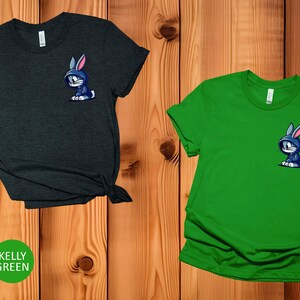 May include: Two t-shirts with a cartoon rabbit wearing a blue hoodie on the pocket. The shirt on the left is heather dark grey and the shirt on the right is kelly green.