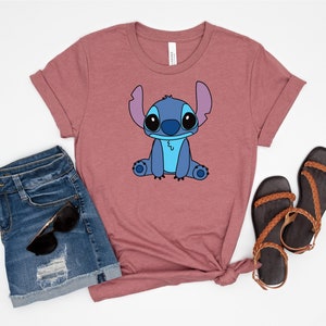 May include: A mauve t-shirt with a cartoon image of Stitch from Lilo & Stitch. The image is in blue, black, and white. The t-shirt is paired with denim shorts and brown sandals.