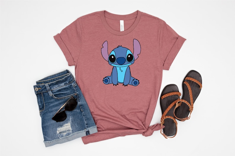 May include: A mauve t-shirt with a cartoon image of Stitch from Lilo & Stitch. The image is in blue, black, and white. The t-shirt is paired with denim shorts and brown sandals.