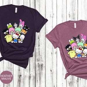 May include: Two t-shirts with a graphic print of a group of cartoon characters. The characters are all different colors and have different expressions. The t-shirts are available in heather maroon and heather mauve.