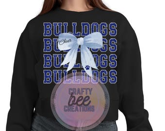 Può includere: Felpa nera con il testo "Bulldogs" ripetuto in glitter blu. Un fiocco blu e bianco con la parola "Cheer" è centrato sulla felpa. Il testo "Crafty Bee Creations" è visibile nell'angolo in basso a destra.