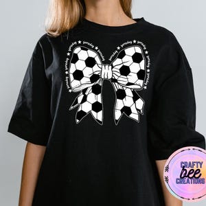 May include: Black t-shirt with a white soccer ball bow design. The bow is made up of soccer balls and has the word "gameday" written around it.