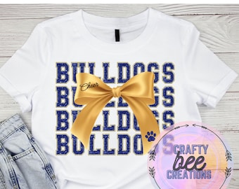 Può includere: T-shirt bianca con la scritta "BULLDOGS" in lettere blu e oro. Un fiocco dorato è centrato sulla maglietta, con la scritta "Cheer" sul fiocco. Un'impronta di zampa è in basso a destra.