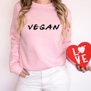 Puede incluir: Una sudadera rosa con el texto "VEGAN" en una fuente estilizada. La persona sostiene una caja roja en forma de coraz&oacute;n con la palabra "LOVE" impresa en ella.