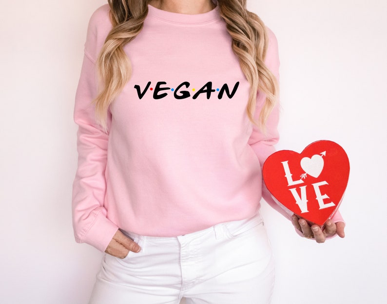 Puede incluir: Una sudadera rosa con el texto "VEGAN" en una fuente estilizada. La persona sostiene una caja roja en forma de coraz&oacute;n con la palabra "LOVE" impresa en ella.