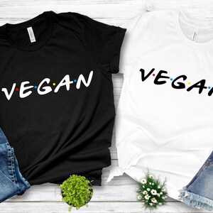 Puede incluir: Dos camisetas con la palabra "VEGAN" escrita en una fuente estilizada, similar al logotipo de "Friends", con puntos de colores entre las letras. Una camiseta es negra y la otra es blanca.