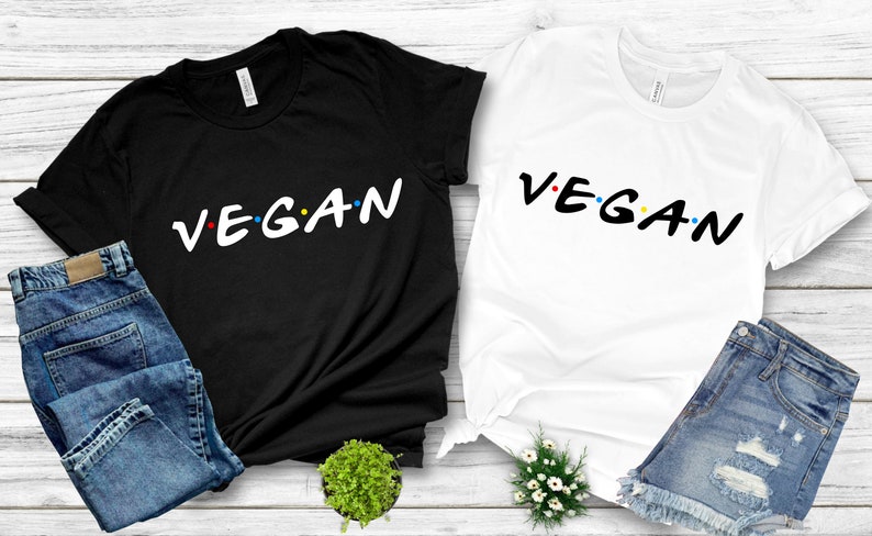 Puede incluir: Dos camisetas con la palabra "VEGAN" escrita en una fuente estilizada, similar al logotipo de "Friends", con puntos de colores entre las letras. Una camiseta es negra y la otra es blanca.