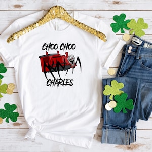 May include: White t-shirt with a red cartoon train with the text "Choo Choo Charles" printed on it. The train has a face with a wide smile and large teeth. The shirt is on a wooden surface with green shamrocks and gold coins.