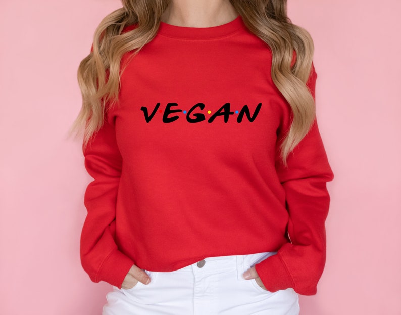 Puede incluir: Sudadera roja con la palabra "VEGAN" escrita en una fuente estilizada, similar al logotipo de la serie "Friends".