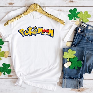 May include: White t-shirt with the word "Pokémon" in a blue and yellow font, with a heart-shaped design. The shirt is paired with blue jeans, shamrock decorations, and gold coins. The shirt is hanging on a gold hanger.