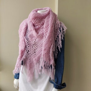 May include: A pink crocheted shawl with fringe on a white mannequin. The shawl is draped around the mannequin's neck and shoulders.