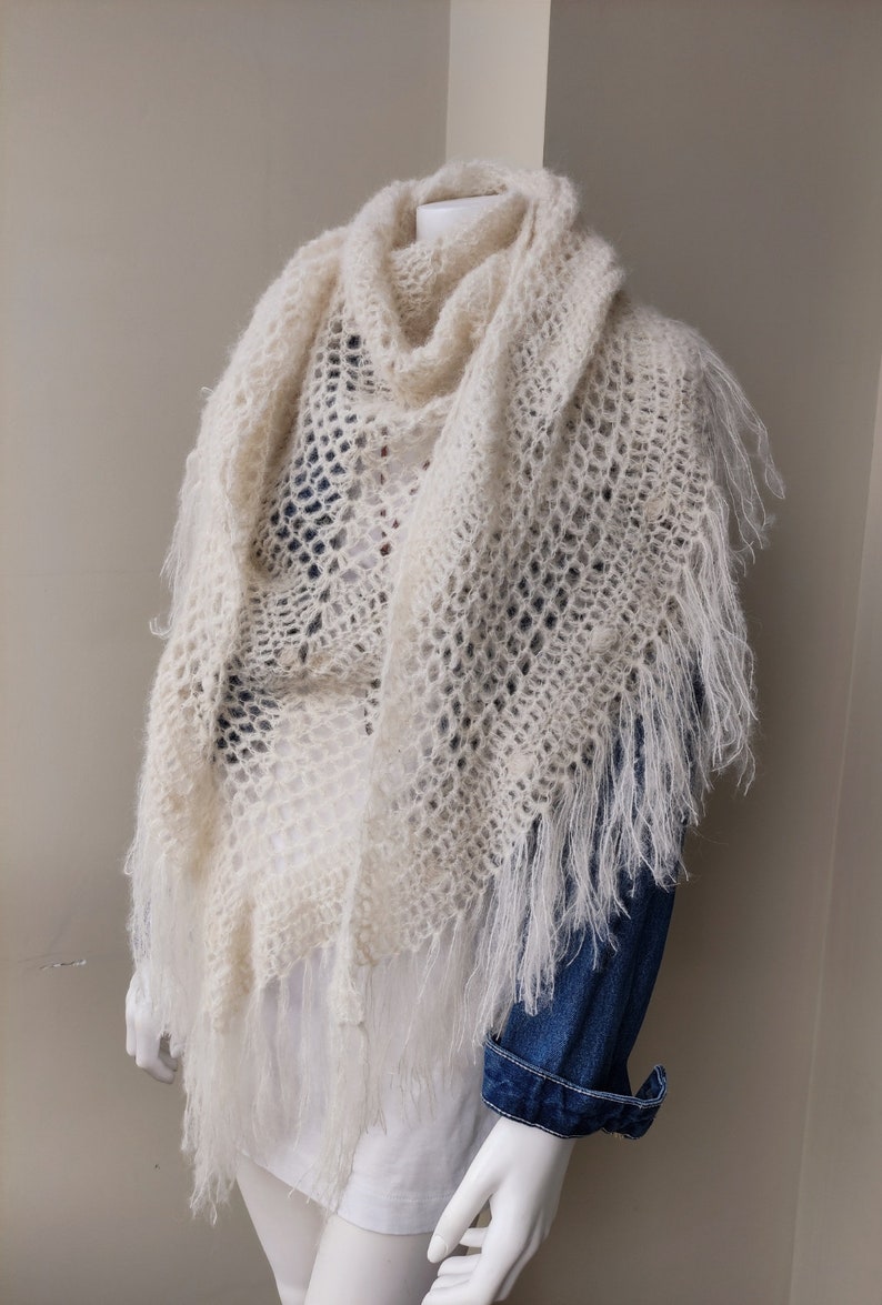 May include: A white crocheted shawl with fringe. The shawl is draped over a mannequin wearing a denim shirt.