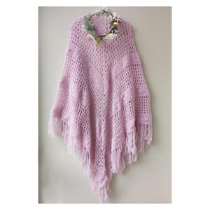 May include: A light purple crochet poncho with fringe detailing. The poncho is hanging on a hanger and has a floral crown on top.