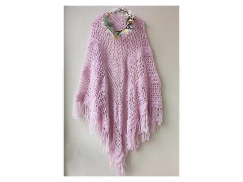 May include: A light purple crochet poncho with fringe detailing. The poncho is hanging on a hanger and has a floral crown on top.
