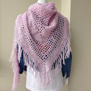May include: A pink crocheted shawl with fringe. The shawl is draped over a mannequin wearing a white t-shirt and a denim jacket.