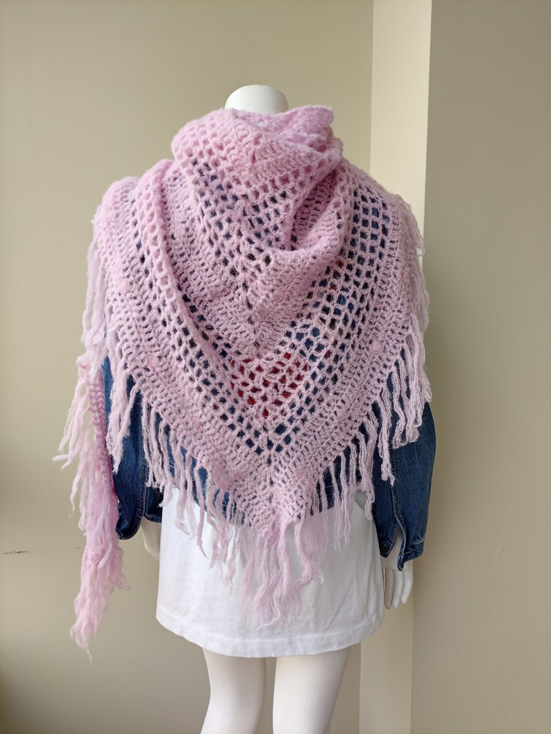 May include: A pink crocheted shawl with fringe. The shawl is draped over a mannequin wearing a white t-shirt and a denim jacket.
