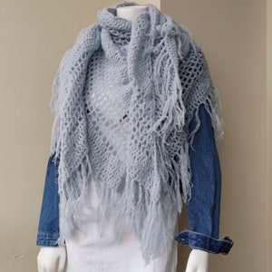 May include: A light blue crocheted shawl with fringe. The shawl is draped over a mannequin wearing a white shirt and a denim jacket.