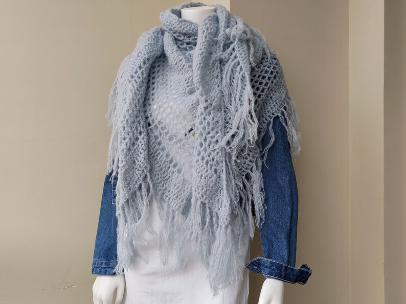 May include: A light blue crocheted shawl with fringe. The shawl is draped over a mannequin wearing a white shirt and a denim jacket.