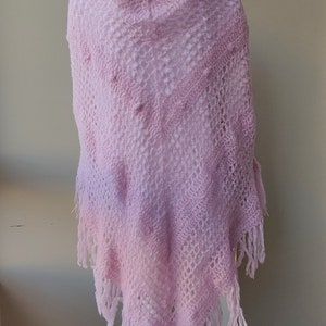 May include: A pink crocheted shawl with a delicate openwork pattern and fringe trim. The shawl is displayed on a white mannequin.