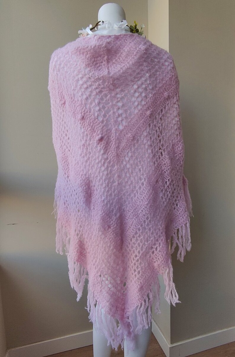 May include: A pink crocheted shawl with a delicate openwork pattern and fringe trim. The shawl is displayed on a white mannequin.