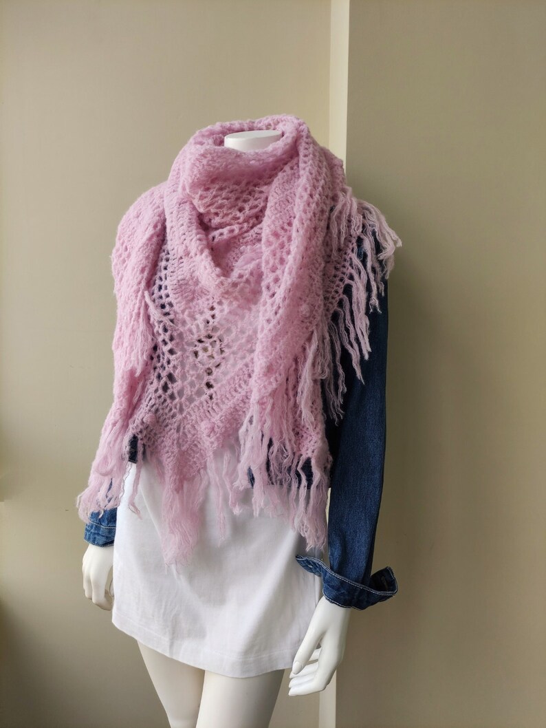May include: A pink crocheted shawl with fringe on a white mannequin. The shawl is draped around the mannequin's neck and shoulders.