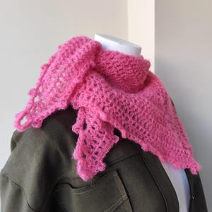 Peut inclure: Écharpe rose vif, crochetée à la main, avec un motif ajouré texturé. L'écharpe est drapée sur un mannequin, mettant en valeur sa forme triangulaire et ses bords décoratifs. L'écharpe est faite d'une laine douce et duveteuse.
