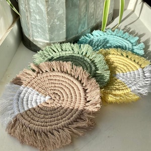 May include: A collection of handmade macrame coasters in various colors, including beige, white, yellow, and teal. Each coaster features a circular design with a fringed edge, perfect for protecting surfaces from drink condensation.