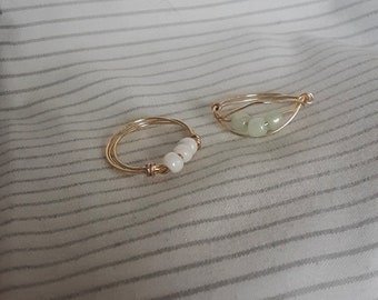 Puede incluir: Dos anillos de alambre hechos a mano. Uno presenta tres cuentas blancas, el otro tres piedras verde pálido. Ambos anillos están hechos con alambre dorado y descansan sobre un fondo de tela a rayas.