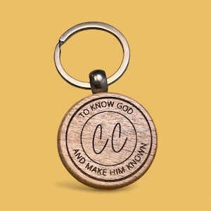 May include: A wooden keychain with a silver key ring. The round wooden pendant has the engraved text "TO KNOW GOD AND MAKE HIM KNOWN" around the edge, with the letters "CC" in the center. The background is a solid yellow.
