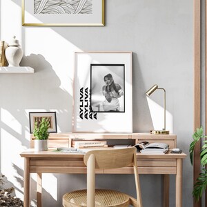 May include: A black and white photo of a woman with a black and white text overlay that says "AYLIVA" repeated multiple times. The photo is framed and hanging on a white wall above a wooden desk with a gold lamp and a potted plant.