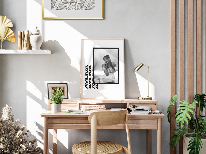 May include: A black and white photo of a woman with a black and white text overlay that says "AYLIVA" repeated multiple times. The photo is framed and hanging on a white wall above a wooden desk with a gold lamp and a potted plant.
