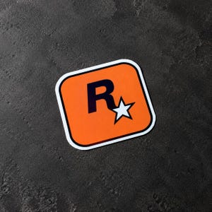 Op de afbeelding: Een vierkante sticker met afgeronde hoeken met een oranje achtergrond, een zwarte letter R en een witte vijfpuntige ster. Het ontwerp is omrand met een dunne witte lijn, wat zorgt voor een contrastrijk grafisch embleem voor diverse oppervlakken.